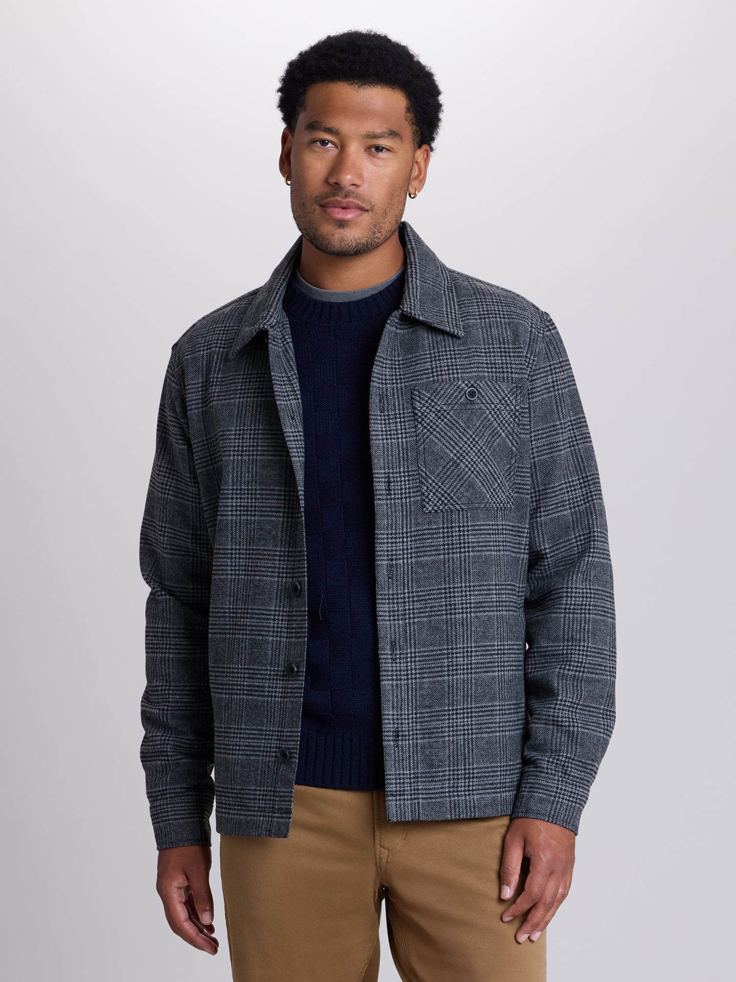 A man wearing a dark gray and navy plaid overshirt jacket over a navy sweater and tan pants, standing against a light gray studio background. | Forged