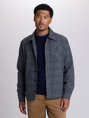 A man wearing a dark gray and navy plaid overshirt jacket over a navy sweater and tan pants, standing against a light gray studio background. | Forged