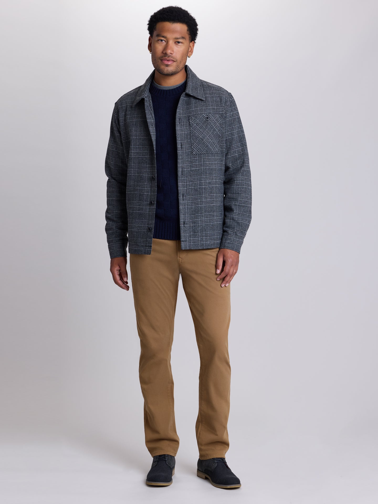 A man modeling a dark gray and blue plaid overshirt over a navy blue sweater, paired with tan pants and dark shoes, against a light gray studio background. | Forged