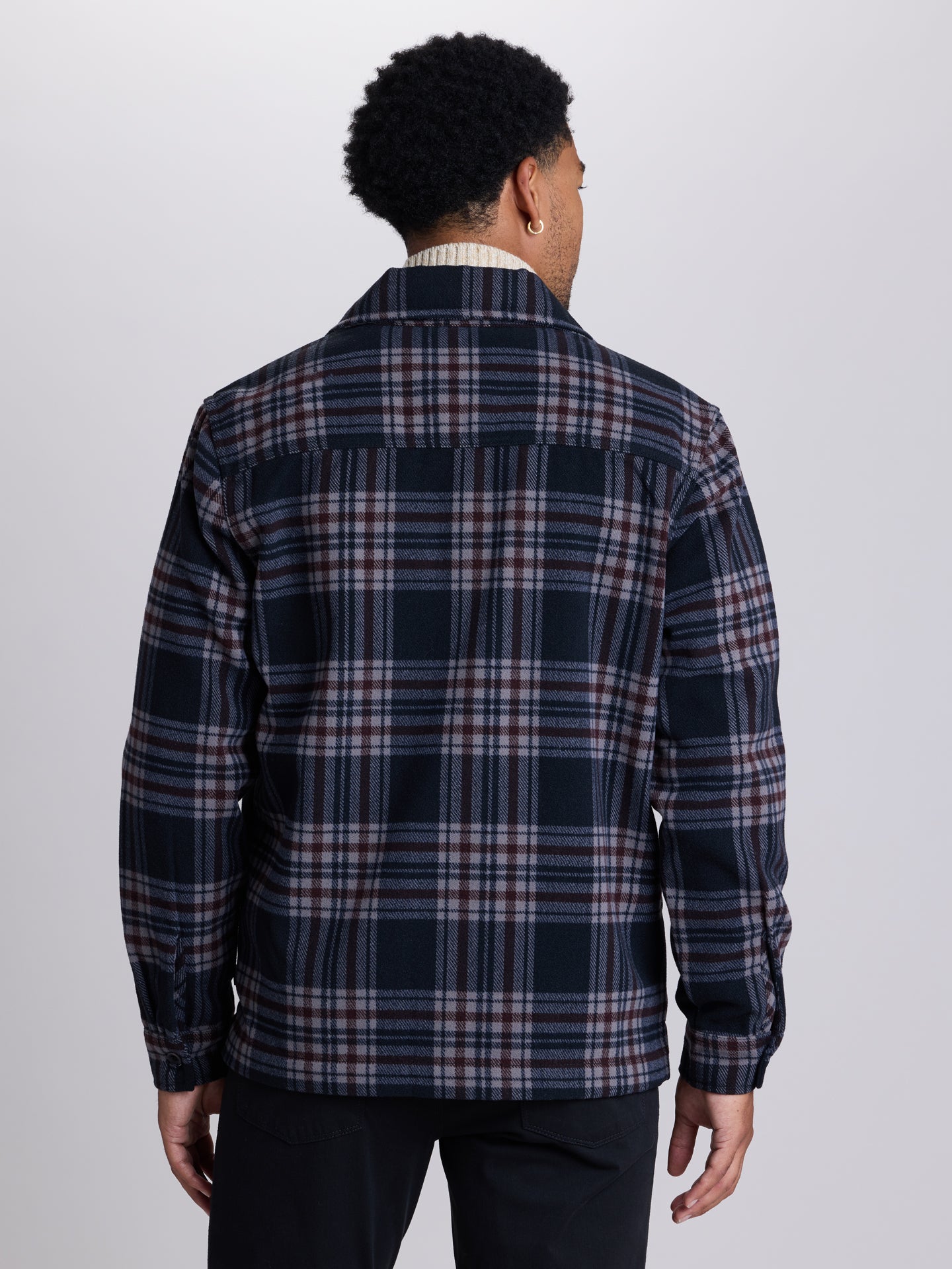 A man viewed from the back wearing a navy blue plaid overshirt with red and white stripes, paired with dark trousers, against a light gray studio background. | Deep Space