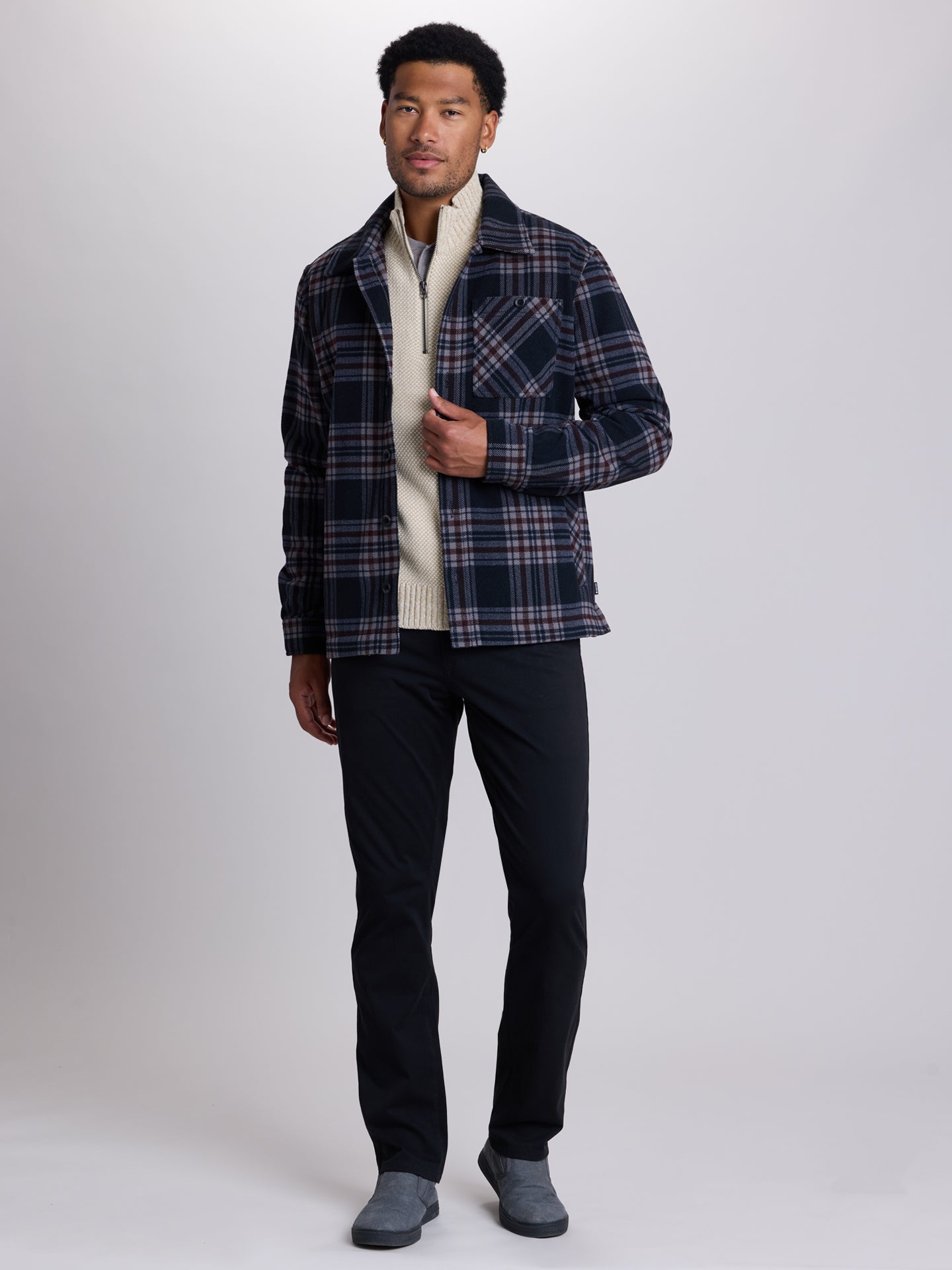 A man modeling a navy blue and black plaid overshirt over a cream knit sweater, paired with black trousers and gray slip-on shoes, against a light gray studio background. | Deep Space