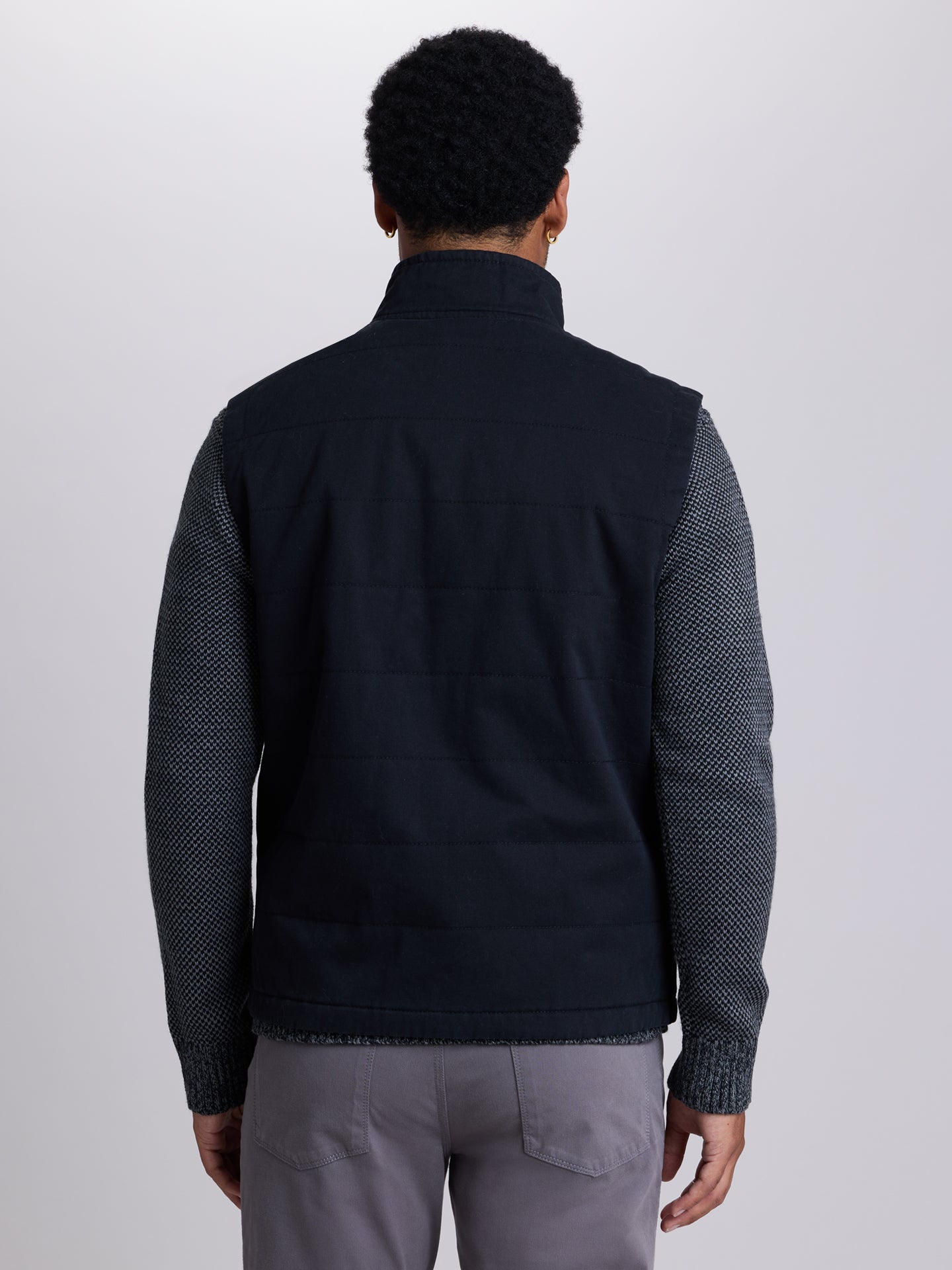 A man viewed from the back wearing a black sleeveless quilted vest over a dark patterned long-sleeve shirt, paired with gray trousers, against a light gray studio background. | Black