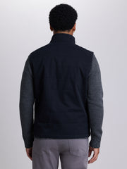 A man viewed from the back wearing a black sleeveless quilted vest over a dark patterned long-sleeve shirt, paired with gray trousers, against a light gray studio background. | Black