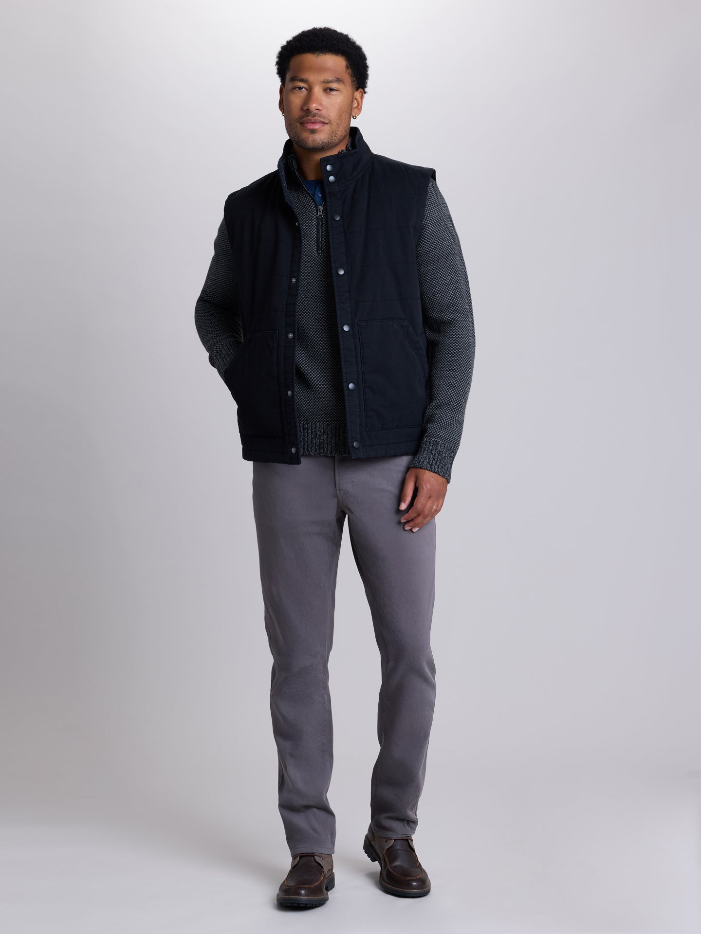 A man modeling a dark navy blue quilted vest with gray knit zip up, paired with gray trousers and brown shoes, against a light gray studio background. | Skillet