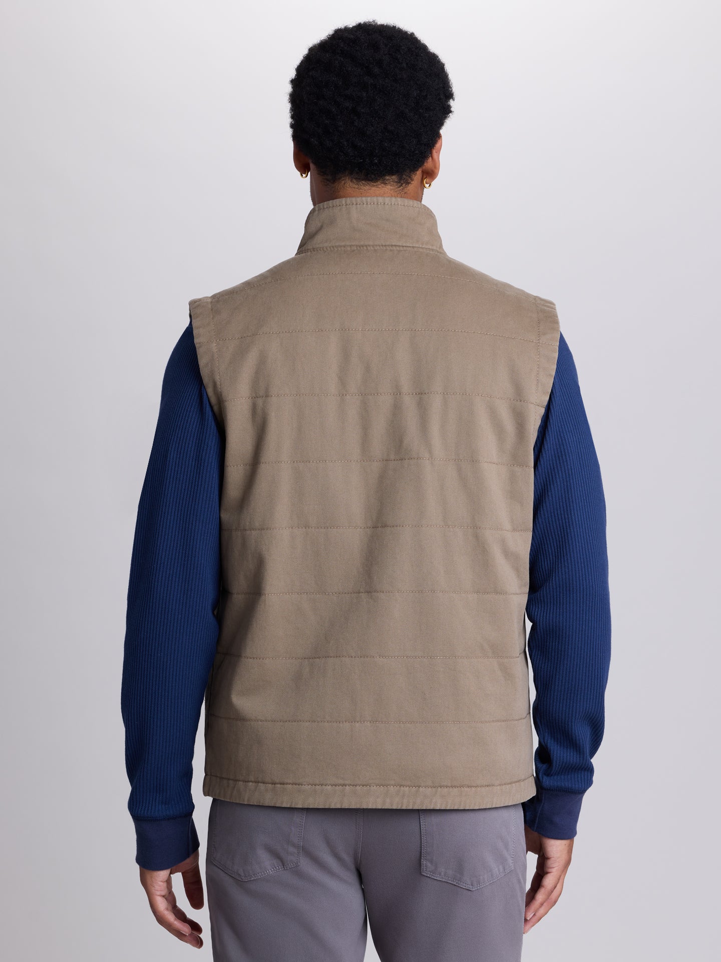 A man viewed from the back wearing a tan quilted puffer vest over a navy blue long-sleeve shirt and gray pants, against a light gray studio background. | Dugout