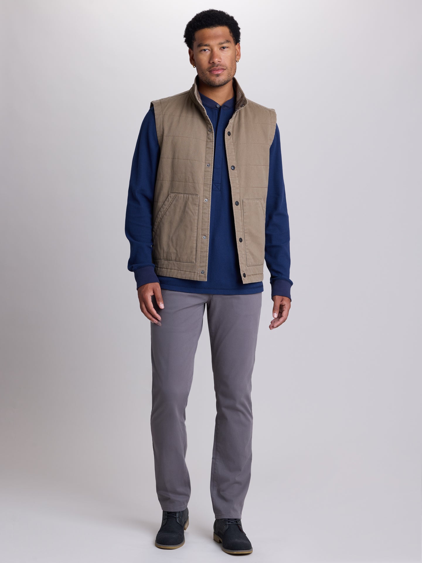 A man modeling a tan quilted sleeveless vest over a navy blue long-sleeve shirt, paired with gray trousers and dark shoes, against a light gray studio background. | Dugout