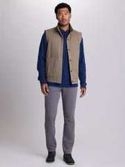 A man modeling a tan quilted sleeveless vest over a navy blue long-sleeve shirt, paired with gray trousers and dark shoes, against a light gray studio background. | Dugout