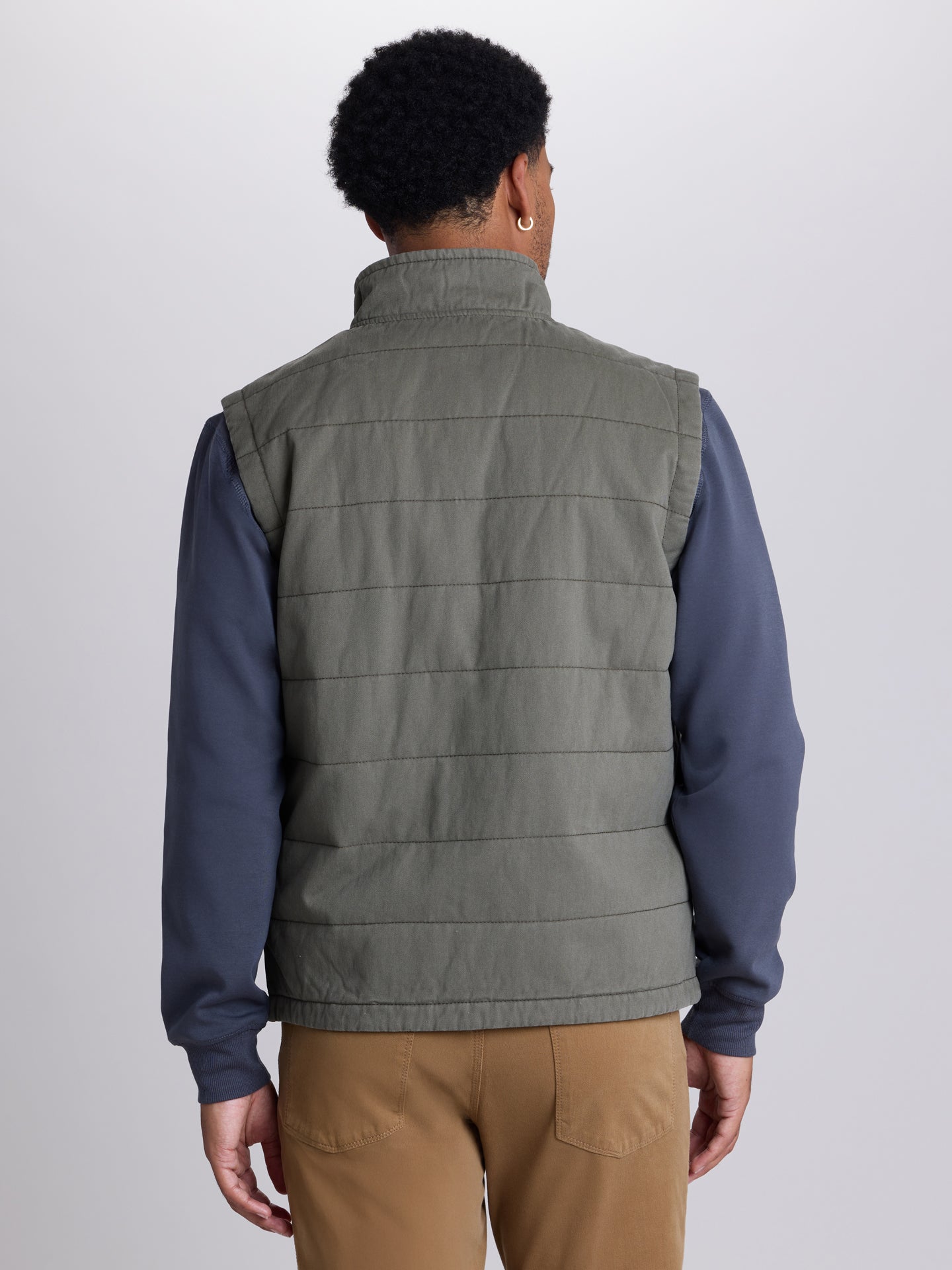 A man viewed from the back wearing a muted olive green quilted puffer vest over a navy blue long-sleeve shirt and tan pants, against a light gray studio background. | Cargo Green