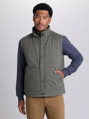 A man wearing a muted olive green quilted puffer vest over a navy blue long-sleeve shirt and tan pants, photographed against a neutral light gray studio background. | Cargo Green