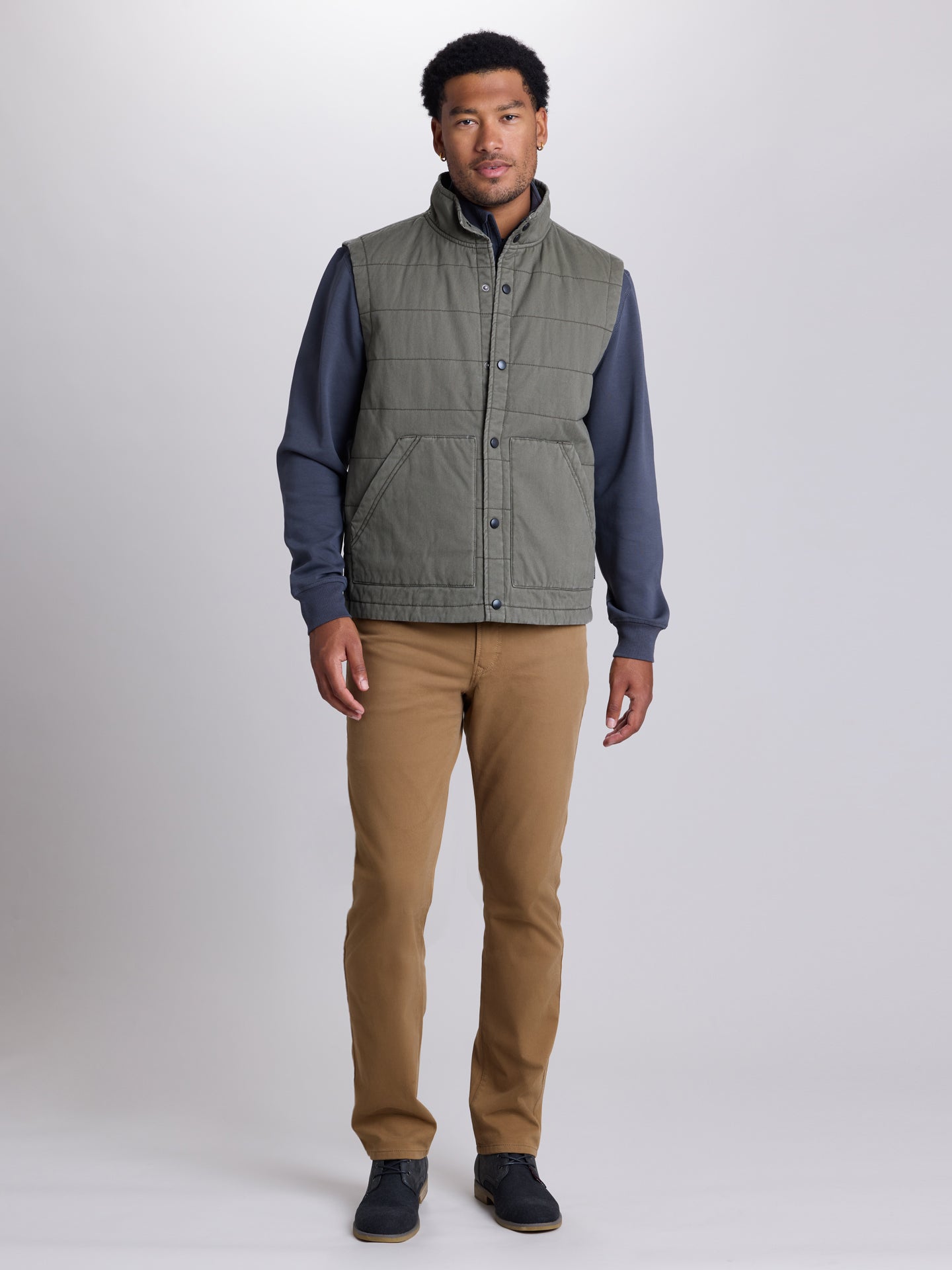 A man modeling a sleeveless quilted vest with a high collar, featuring a muted sage green body and navy blue sleeves, paired with tan pants and dark shoes, against a light gray studio background. | Cargo Green