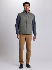 A man modeling a sleeveless quilted vest with a high collar, featuring a muted sage green body and navy blue sleeves, paired with tan pants and dark shoes, against a light gray studio background. | Cargo Green